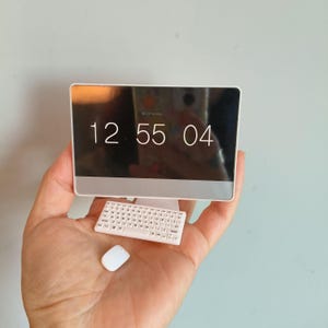 Op de afbeelding: Een miniatuurcomputer met een witte lijst, met de tijd 12:55:04 op het scherm. Een klein wit toetsenbord en muis bevinden zich onder het scherm. De computer wordt in een hand gehouden.
