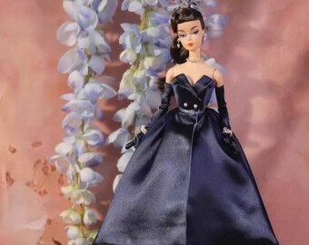 May include: A doll in a navy blue satin gown with a sweetheart neckline, long gloves, and a matching headpiece. The dress has a full skirt and a silver belt at the waist. The doll is positioned in front of a backdrop of light blue flowers.
