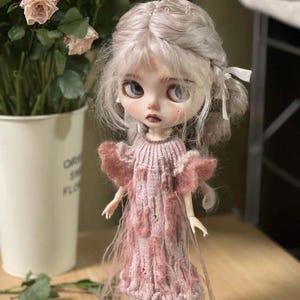 May include: A collectible doll with pale skin, gray hair, and large eyes. The doll wears a pink knitted dress with fringed accents. The doll is standing on a wooden base, with a blurred background of flowers in a white pot.