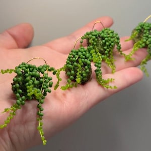 May include: Miniature green plant ornaments, resembling string of pearls, are held in a hand. Each ornament features a spherical base covered in small, round green beads, with delicate, trailing green stems. The ornaments have small gold wire loops for hanging.