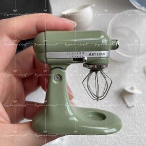 Peut inclure: Un mini-batteur sur socle Artisan vert sauge avec un fouet en fil de fer. Le batteur a un cadran avec des chiffres et le mot "ARTISAN" imprimé sur le côté. Le batteur est une petite réplique d'un appareil de cuisine.