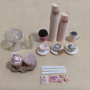 Könnte beinhalten: Eine Sammlung von Miniatur-Kaffee-Shop-Artikeln, darunter gestapelte rosa Pappbecher, kleine Kaffeetassen mit Untertassen und zwei Mixgetränke mit Schlagsahne. Weitere Artikel sind ein Milchkännchen, Zuckerpäckchen und Rührstäbchen.