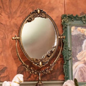 Op de afbeelding: Een ovale make-upspiegel in antieke stijl met een bronzen afwerking. De spiegel heeft sierlijke bloemendetails en een kralenrand. Hij staat op een decoratieve voet en heeft verstelbare armen. Een wit schommelpaard en een roze roos staan op de voorgrond.