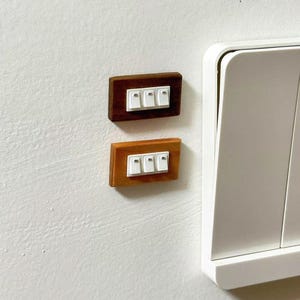 May include: Two wooden light switch covers, one dark brown and one light brown, each with three white switches. The covers are mounted on a white wall, next to a larger white light switch.