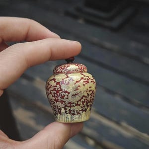 May include: A small, decorative jar with a lid, displaying a red and gold pattern. The jar's surface is adorned with detailed designs, possibly including a dragon. The jar is held in a hand, highlighting its size and the intricacy of the design.