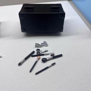 May include: A black miniature suitcase with silver hardware is displayed with several makeup brushes and tools. The brushes have black handles and various brush heads. The tools include small scissors and other silver accessories, all arranged on a white surface.