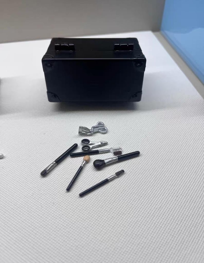 May include: A black miniature suitcase with silver hardware is displayed with several makeup brushes and tools. The brushes have black handles and various brush heads. The tools include small scissors and other silver accessories, all arranged on a white surface.