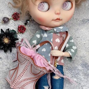 May include: A doll with large, detailed eyes and rosy cheeks, wearing a patchwork top with heart patterns and distressed denim jeans. The doll carries a pink purse with a bow and has a matching pink and brown strap. Accessories include rose earrings.