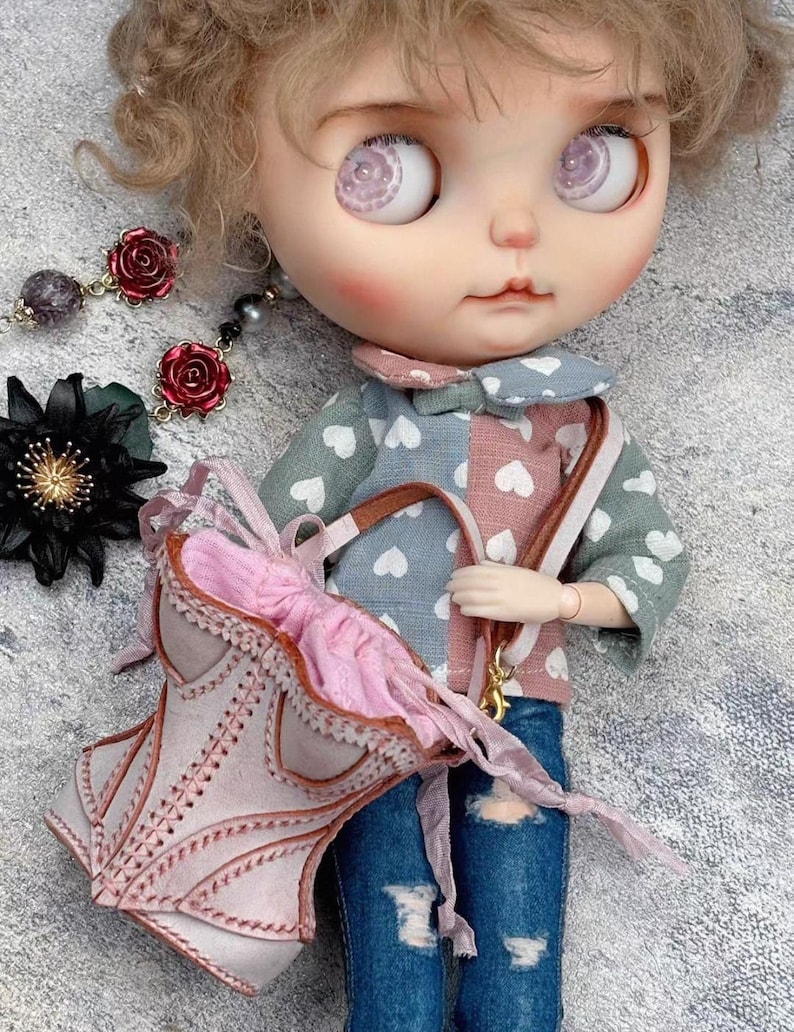 May include: A doll with large, detailed eyes and rosy cheeks, wearing a patchwork top with heart patterns and distressed denim jeans. The doll carries a pink purse with a bow and has a matching pink and brown strap. Accessories include rose earrings.