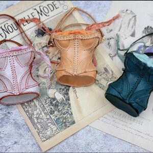 May include: Three miniature corset-shaped handbags in pastel colors: pink, tan, and teal. Each bag has a handle and decorative stitching, resembling vintage corsets. They rest on a vintage newspaper with the text "LA NOUVELLE MODE".