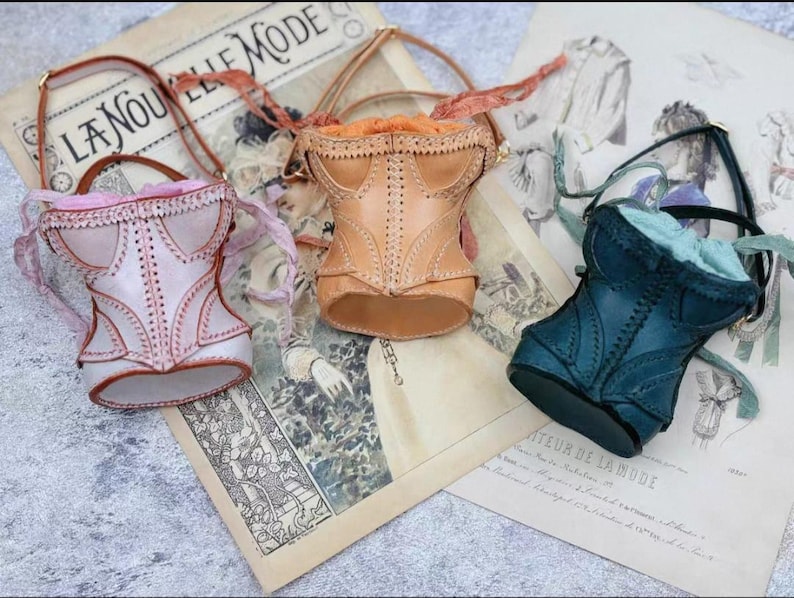May include: Three miniature corset-shaped handbags in pastel colors: pink, tan, and teal. Each bag has a handle and decorative stitching, resembling vintage corsets. They rest on a vintage newspaper with the text "LA NOUVELLE MODE".