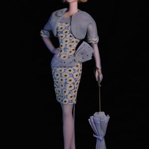 May include: A vintage doll wearing a light blue floral dress, a matching bolero jacket, and a wide-brimmed hat. The doll holds a closed, light blue umbrella. The doll is wearing white heels and sheer stockings.