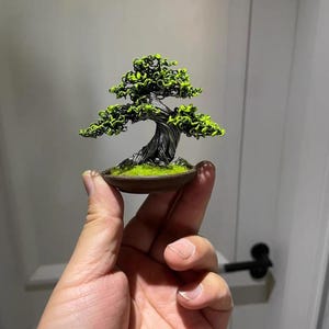 Könnte beinhalten: Ein Miniatur-Bonsai-Baum mit einem verdrehten, dunkelgrauen Stamm und leuchtend grünem Laub. Der Baum steht in einem kleinen, braunen Keramiktopf mit grünem Moos. Der Bonsai wird in der Hand einer Person gehalten und zeigt seine geringe Größe.