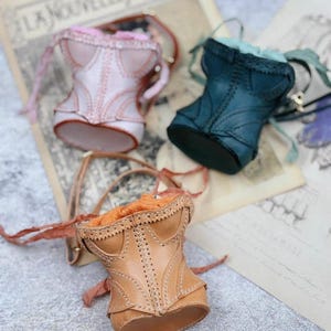 May include: Three miniature corset-shaped bags in pink, teal, and tan leather. Each bag features intricate stitching and a drawstring closure. The bags are displayed on a vintage paper background, evoking a sense of classic fashion.