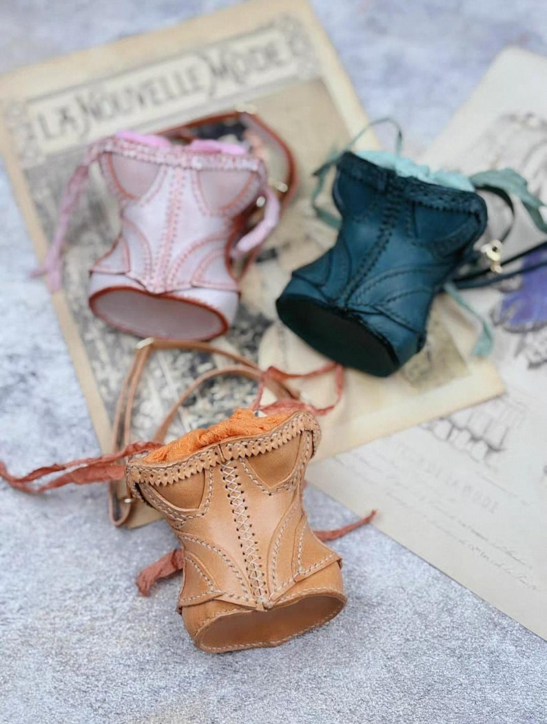 May include: Three miniature corset-shaped bags in pink, teal, and tan leather. Each bag features intricate stitching and a drawstring closure. The bags are displayed on a vintage paper background, evoking a sense of classic fashion.