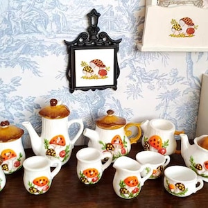 Op de afbeelding: Een verzameling keramische keukenspullen met een grillig paddestoelontwerp. De set bevat theepotten, kopjes, suikerpotten en een kleine kast. Het kleurenpalet is voornamelijk wit, bruin, oranje en groen, met een gedetailleerde paddestoelillustratie.