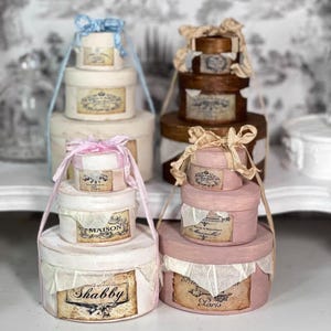 Puede incluir: Cajas redondas decorativas apiladas en crema, marrón y rosa, adornadas con lazos y etiquetas de estilo vintage. Cada juego tiene un asa. Las cajas están etiquetadas como "Shabby", "Maison" y "Paris".