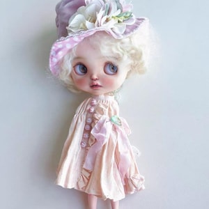 Puede incluir: Muñeca hecha a mano con un vestido rosa pálido, adornada con botones rosas y un lazo grande. La muñeca lleva un sombrero lavanda con flores blancas y un borde de lunares rosas. Los zapatos de la muñeca son negros con suelas marrones.