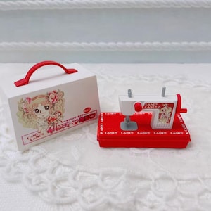 May include: A miniature sewing machine and a white box with a red handle. The sewing machine is white and red, with the words "Candy Candy" on it. The box has a cartoon illustration of a girl.
