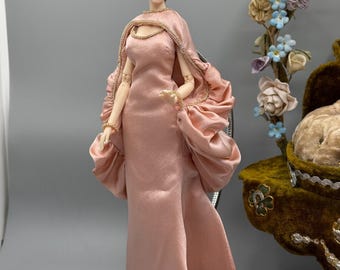 May include: A fashion doll wearing a pink satin gown with a matching cape and ruffled sleeves. The doll has brown hair and is accessorized with gold jewelry. The doll is standing in front of a gray background.