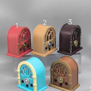 May include: A selection of miniature vintage-style radios in different colours. The radios feature a classic arched design with speaker grilles, dials, and knobs. Colours include red, tan, brown, turquoise, and brown with gold. The radios are displayed on a clear surface.