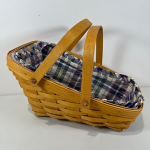 Peut inclure: Un panier rectangulaire en bois tressé doté de deux hautes anses arquées. Le panier présente une finition brun clair et est doublé d'un tissu à motif écossais vert foncé, bleu marine et blanc. La structure est assemblée par de petits rivets.