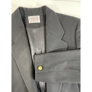 Pode incluir: Um blazer preto com lapela entalhada e um único botão dourado na manga. O blazer tem uma etiqueta dentro da gola. O tecido parece ser uma mistura de lã.