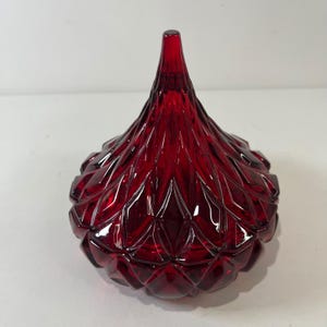 Puede incluir: Un tarro de vidrio rojo rubí con una parte superior puntiaguda y un exterior con diseño de diamantes. El tarro es una pieza decorativa, probablemente para guardar objetos pequeños o como objeto de exhibición. El tarro es de vidrio.