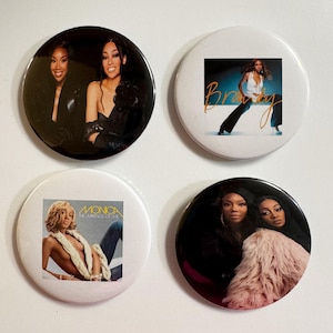 May include: Four round pinback buttons featuring images of female singers. Two buttons have images of two women, one in black and the other in a pink fur coat. One button has the word "Brandy" in orange script. The fourth button has the words "Monica" and "The Boy Is Mine".