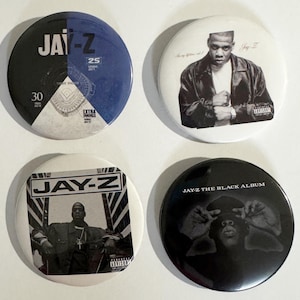 May include: Four round buttons featuring images of Jay-Z. One button has a blue and black design with text, including "JAY-Z" and "25". Another shows Jay-Z in a black and white photo. The third button has a black and white image of Jay-Z with the text "JAY-Z". The last button is black with the text "JAY-Z THE BLACK ALBUM".