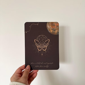 Op de afbeelding: Een donkerbruine kaart met een gouden vlinderontwerp, een halve maan en een planeetillustratie. De kaart heeft de tekst "She's a 10 but she can't separate fiction from reality."