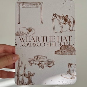 以下が含まれることがあります： カウボーイをテーマにした要素の茶色のイラストが描かれた長方形のカード。牧場の入り口、鞍をつけた馬、カウボーイハット、納屋、ピックアップトラック、サボテン、カウボーイブーツが含まれています。「WEAR THE HAT RIDE THE COWBOY」と書かれています。