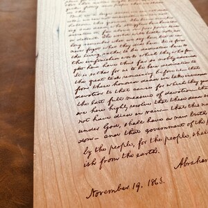 May include: A close-up of a wooden plaque with a handwritten message in brown ink. The message is a quote from Abraham Lincoln's Gettysburg Address, dated November 19, 1863. The quote reads: "It is rather for us to be here dedicated to the great task remaining before us - that from these honored dead we take increased devotion to that cause for which they gave the last full measure of devotion - that we here highly resolve that these dead shall not have died in vain - that this nation, under God, shall have a new birth of freedom - and that government of the people, by the people, for the people, shall not perish from the earth."
