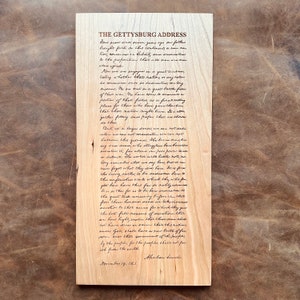 May include: A close-up of a wooden plaque with the Gettysburg Address handwritten in black ink. The text is centered on the plaque and includes the date "November 19, 1863" and the signature "Abraham Lincoln".