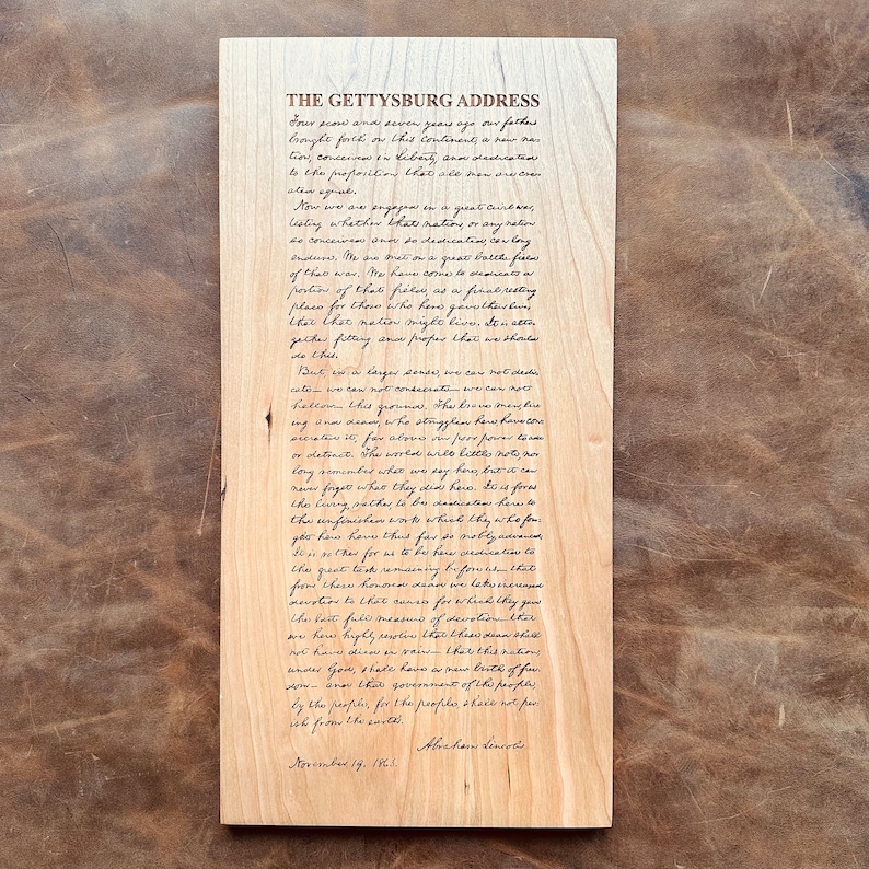 May include: A close-up of a wooden plaque with the Gettysburg Address handwritten in black ink. The text is centered on the plaque and includes the date "November 19, 1863" and the signature "Abraham Lincoln".