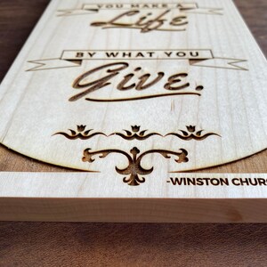 Op de afbeelding: Een houten bord met de quote "Je maakt een leven door wat je geeft." - Winston Churchill. De quote is in het hout gegraveerd en de achtergrond is lichtbruin.