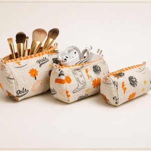 May include: Three cream-colored fabric storage pouches with orange and black patterns. The pouches are filled with makeup brushes, earbuds, and cords. The pouches have the word "Vols" and a "T" logo.