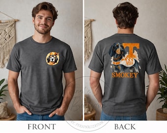 May include: A dark gray t-shirt featuring a small circular graphic on the front chest of a dog wearing sunglasses and a bandana. The back displays a large graphic of a dog wearing a jersey with the text "VOLS" and "SMOKEY" below it.