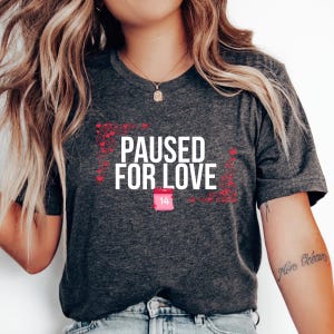 May include: Dark grey t-shirt featuring the white text "PAUSED FOR LOVE", red heart decorations, and a calendar icon with the number 14. A Valentine's Day themed graphic tee.