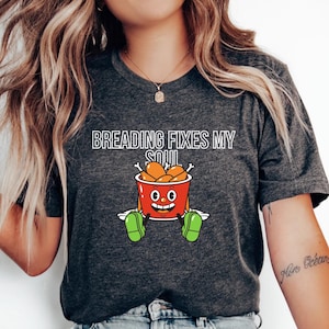 May include: Dark grey t-shirt featuring the text "Breading Fixes My Soul" above a cartoon bucket of fried chicken. The bucket has a smiling face and green shoes. The shirt is a casual style.
