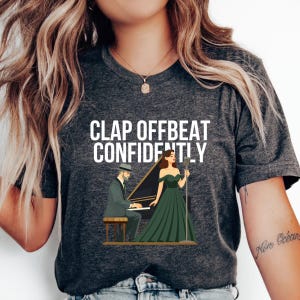 May include: Dark gray t-shirt with the text "CLAP OFFBEAT CONFIDENTLY". The graphic shows a man playing a piano and a woman singing into a microphone, both in front of the piano. The woman is wearing a green dress.