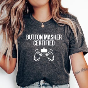 May include: Dark gray t-shirt with the text "BUTTON MASHER CERTIFIED" above a white graphic of a video game controller. The shirt is a crew neck style.