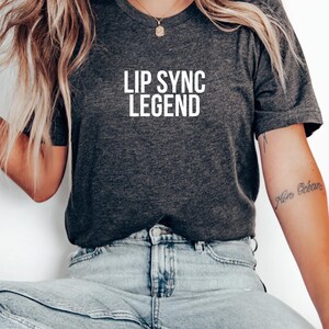 May include: Dark gray t-shirt with the text "LIP SYNC LEGEND" in bold white letters. The shirt is a crew neck style and appears to be made of a soft material. The model is wearing blue jeans.