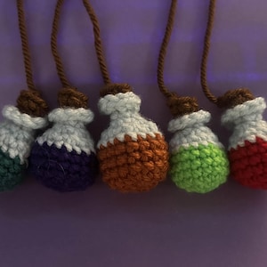 May include: Five crocheted potion bottle ornaments in a row. Each bottle has a different color: green, purple, orange, green, and red. The bottles have white and brown accents and are suspended by brown cords.