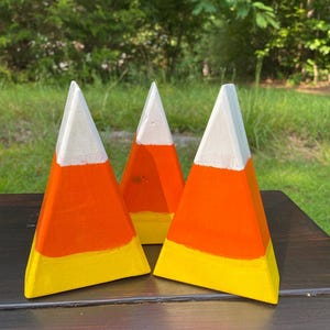 May include: Three wooden candy corn decorations. Each piece is painted with a white tip, orange middle, and yellow base. The decorations are arranged on a dark surface with a green background.