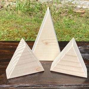 May include: Three wooden pyramid sculptures of varying sizes. The pyramids are made of light-colored wood and have a natural finish. The pyramids are arranged on a dark wooden surface, with a grassy background.