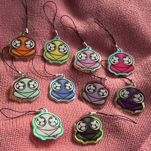 May include: A collection of colorful frog-shaped charms with black cords. Each charm features a different color scheme, including red, orange, yellow, pink, green, blue, purple, and black. The charms have a cartoonish design with large eyes.