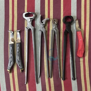 Puede incluir: Una colección de herramientas de herrador, que incluye tenazas, cuchillos para cascos y un cuchillo para recortar cascos. Las herramientas están hechas de metal y madera, con mangos plateados, negros y rojos. Dispuestas sobre un fondo a rayas.