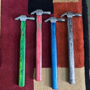 Peut inclure: Quatre marteaux en métal avec des manches en bois colorés. Les manches sont teints en vert, rouge, bleu et gris. Les têtes de marteau sont argentées. Les marteaux sont disposés verticalement sur un tapis à motifs.