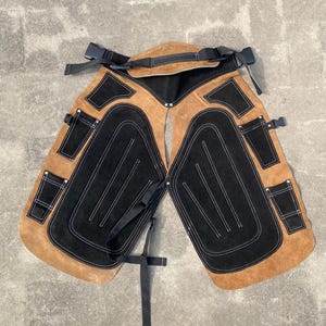 Peut inclure: Chaps de travail en cuir marron et noir avec des coutures blanches. Les chaps ont plusieurs poches et des sangles réglables. Les chaps sont conçus pour protéger les jambes et la taille pendant le travail.